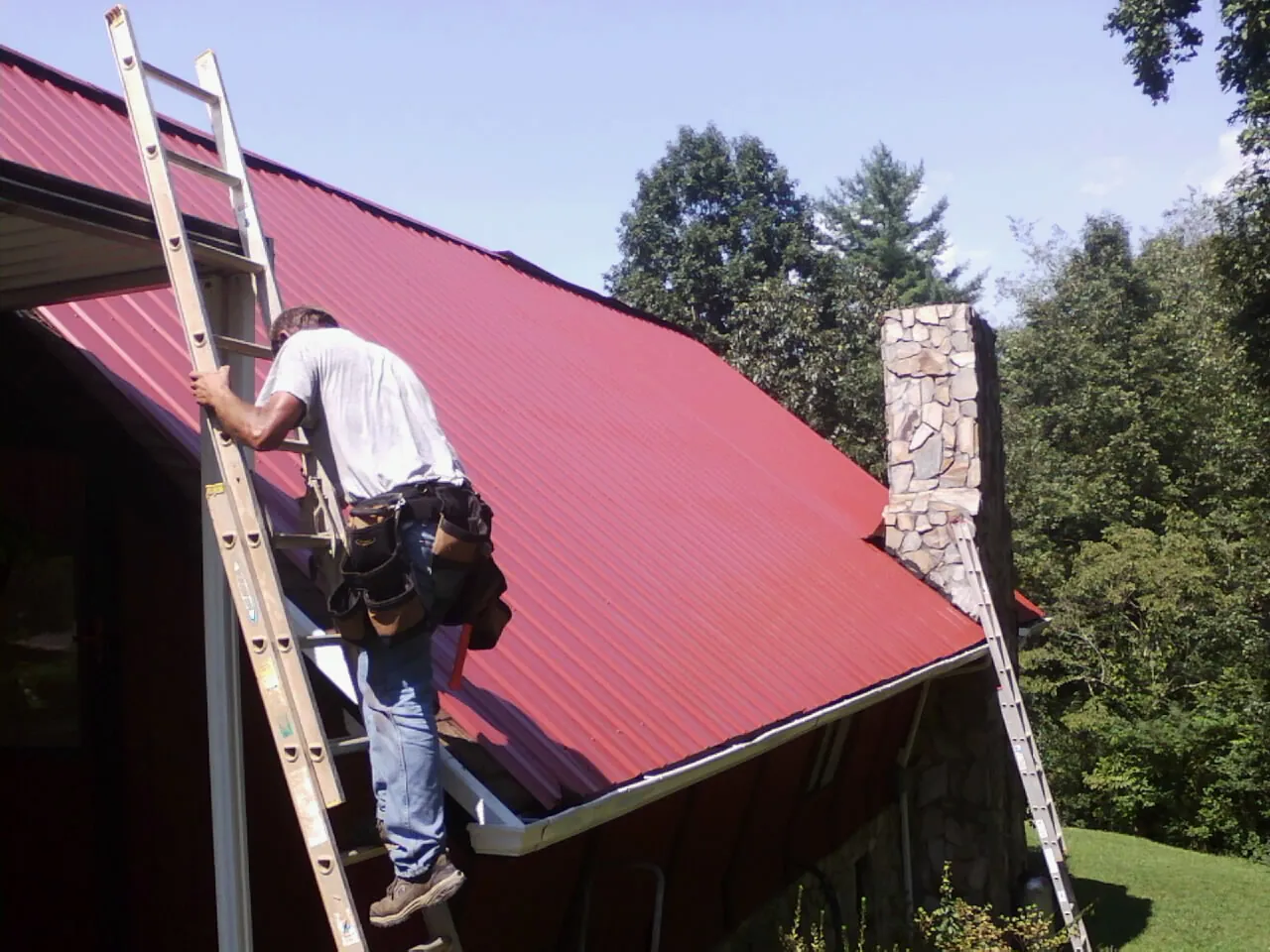 Roofer on a ladder beside a red metal roof and stone chimney — Soffit & Fascia Repair by Roofing Brothers in Fruitport